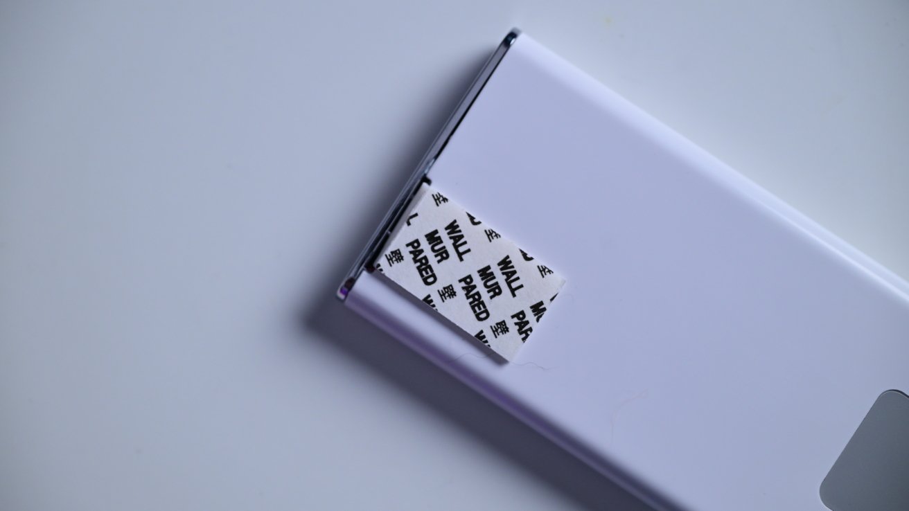 Partially opened white foldable device, revealing a piece of adhesive tape with multilingual text including 'WALL' and 'PARED'.