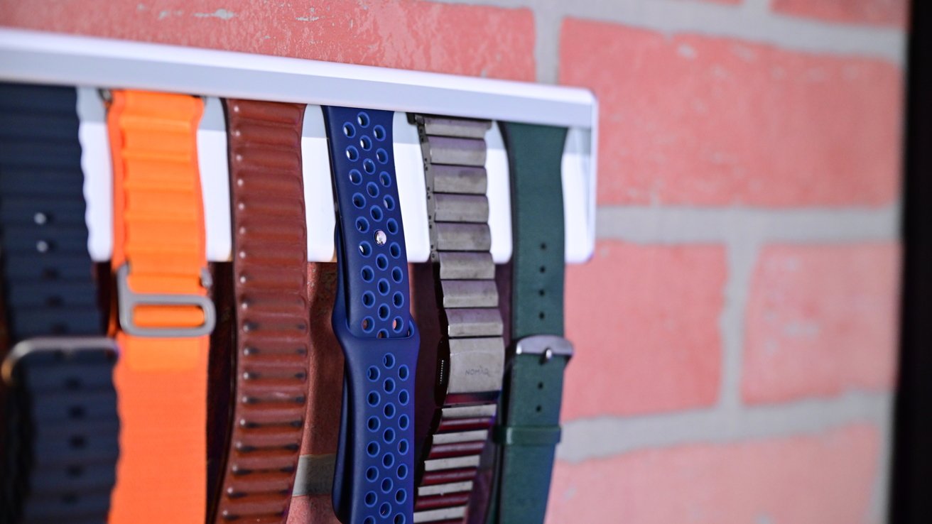 Various colorful watch bands hanging on a wall-mounted metal rack against a brick wall background.