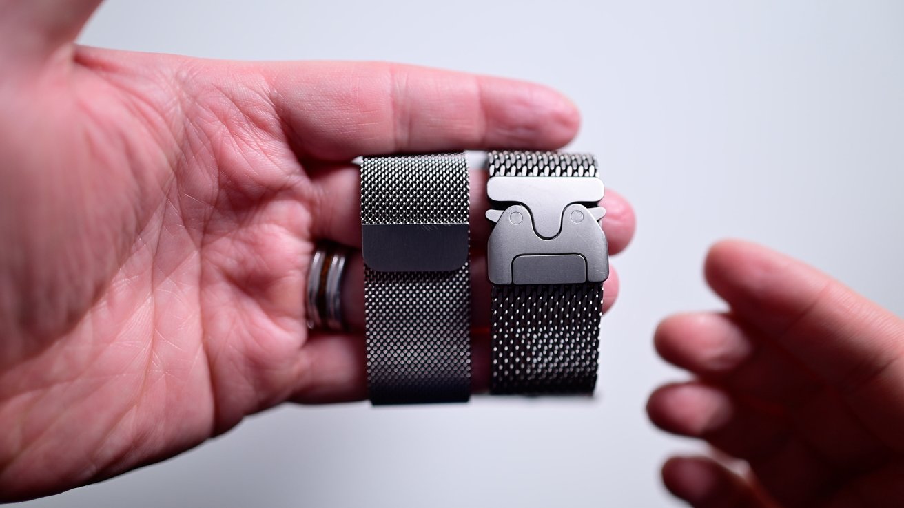 Two hands holding a pair of silver mesh watch straps with metallic clasps against a light background.