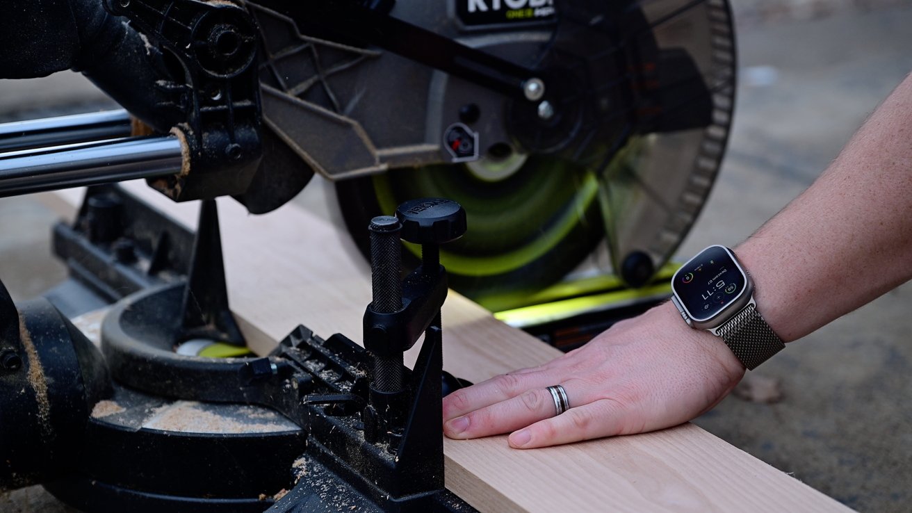 Person's hand wearing a smartwatch, guiding a piece of wood through a miter saw with a rotating blade.