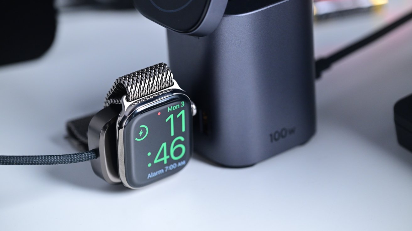 Smartwatch with mesh band charging on a gray 100W charger, displaying the time 11:46 and an alarm set for 7:00 AM.