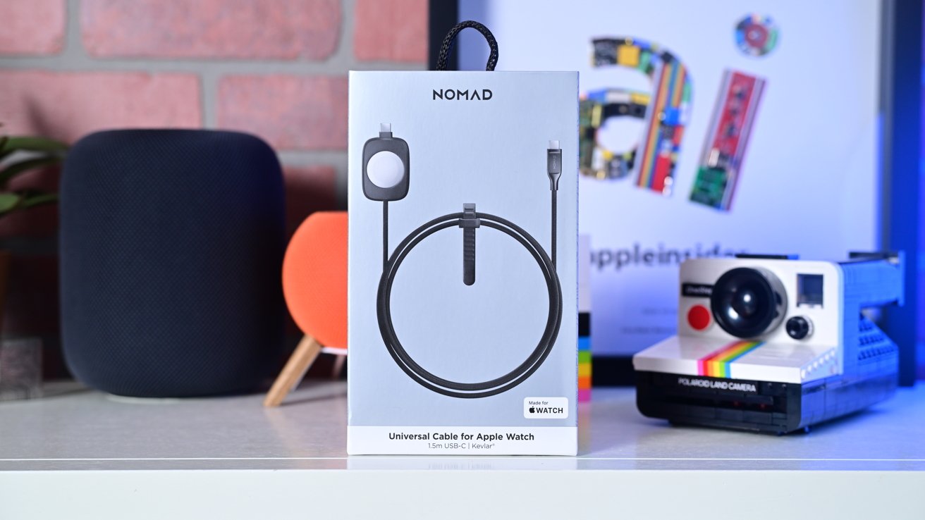 Box of a Nomad universal cable for Apple Watch, surrounded by a speaker, an orange device, and a vintage Polaroid camera on a shelf.