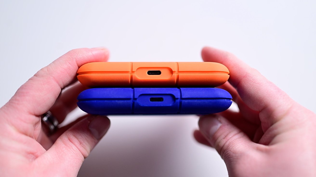 Two hands holding an orange and a blue portable hard drive horizontally, showing USB-C ports on a white background.