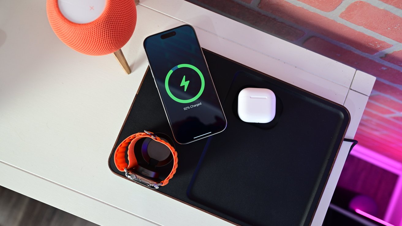 A smartphone charging wirelessly on a pad, displaying 92% charge, next to wireless earbuds and an orange-banded smartwatch, with a smart speaker nearby.