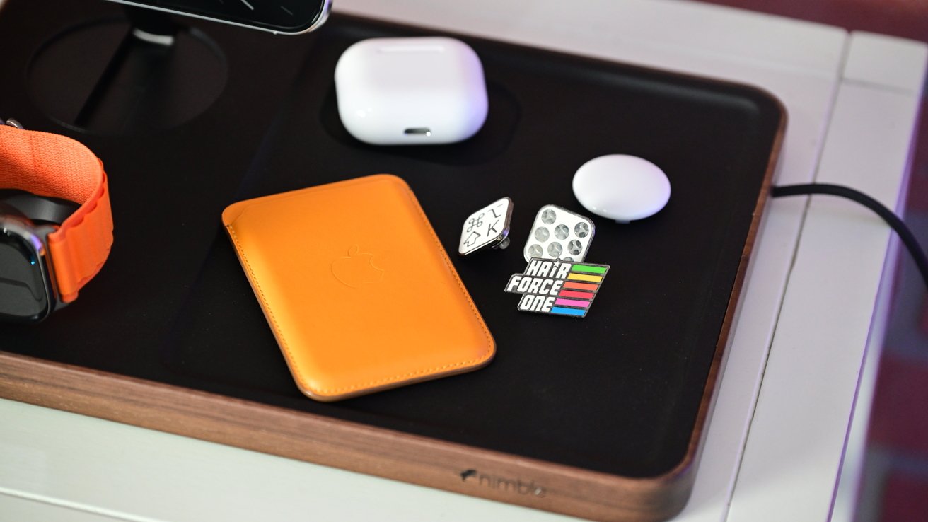 A charging station with an orange wallet, smartwatch, AirPods case, circular pad, and colorful pins, including 'Hair Force One' text.